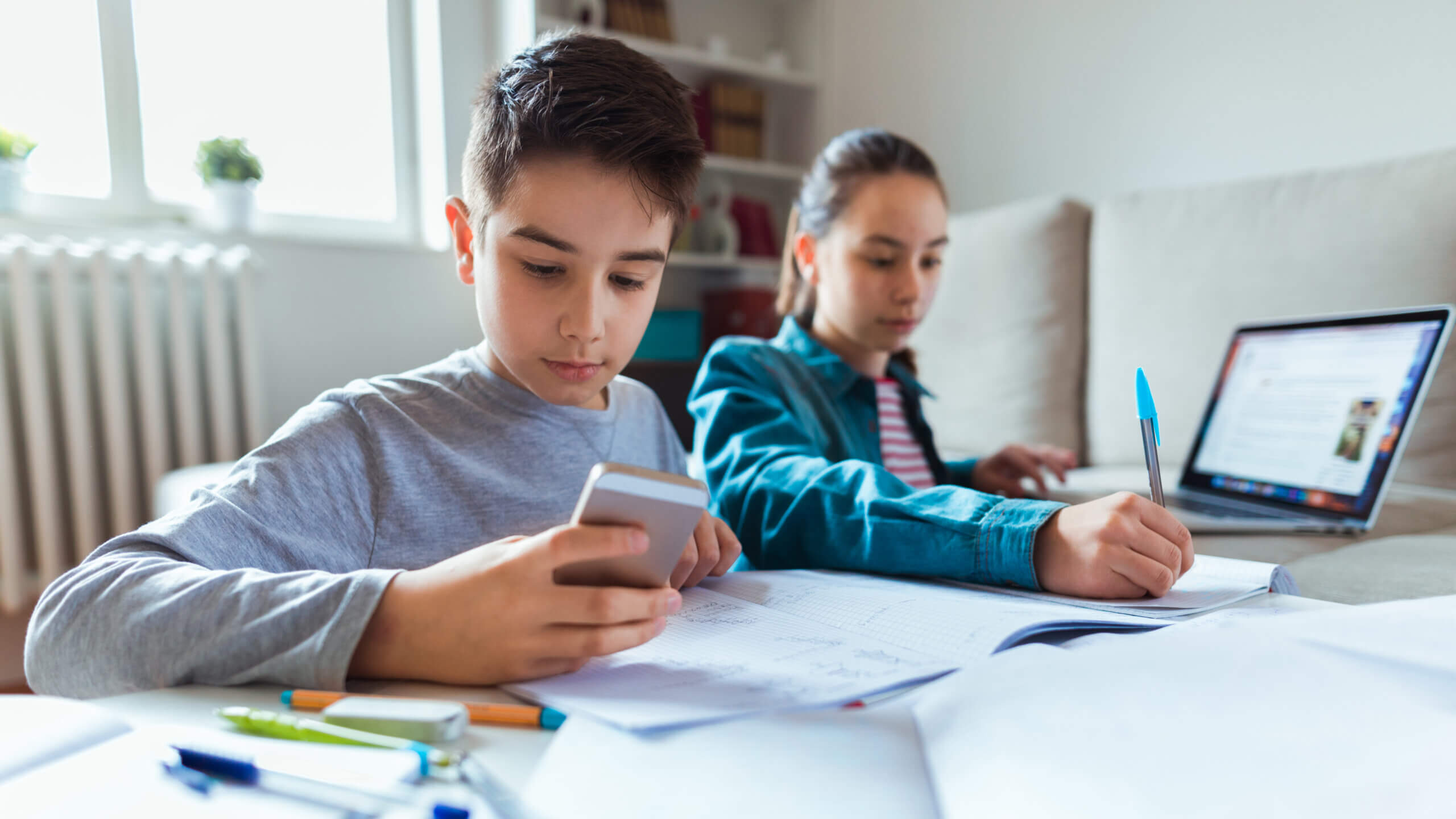 A boy and girl use a laptop and smartphone to do their homework